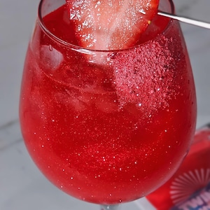 May include: A bright red cocktail in a stemmed glass, garnished with a heart-shaped strawberry slice. The drink has a glittery appearance, and a metal skewer holds the garnish. The glass is on a light surface. The drink is likely a cocktail.