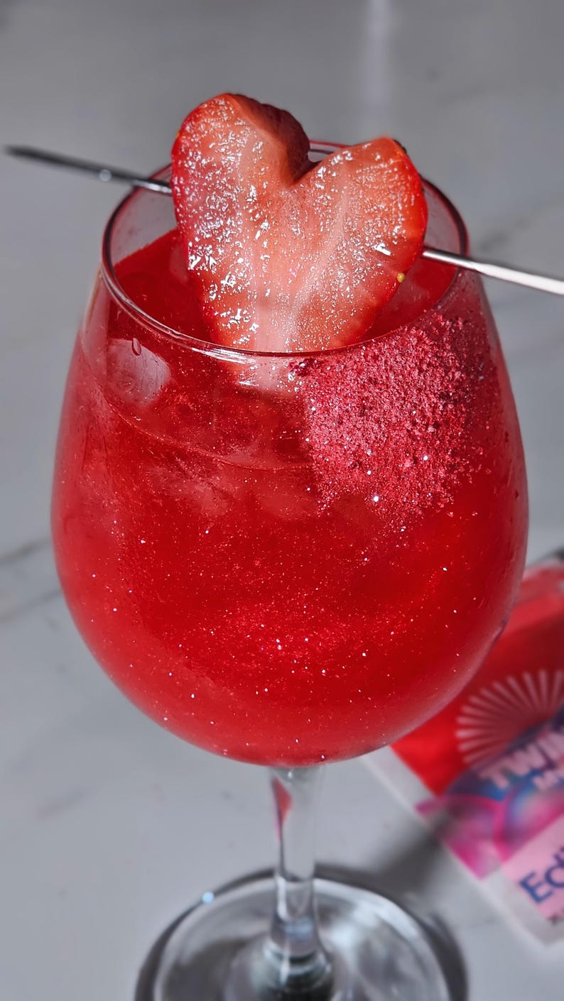 May include: A bright red cocktail in a stemmed glass, garnished with a heart-shaped strawberry slice. The drink has a glittery appearance, and a metal skewer holds the garnish. The glass is on a light surface. The drink is likely a cocktail.