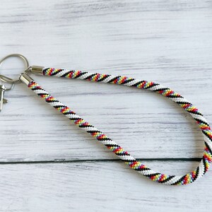 May include: A white beaded keychain with black, red, yellow, and blue stripes. The keychain has a silver key ring and a silver clasp.
