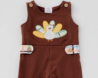 May include: Brown baby overalls with a turkey applique on the front. The overalls have a plaid trim and snap closures on the legs.