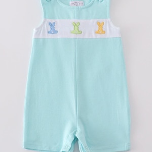 Puede incluir: Un pelele de bebé azul claro con una banda blanca en el pecho que presenta tres conejos bordados en diferentes colores. El pelele tiene un diseño sin mangas y cierres a presión en la parte inferior.