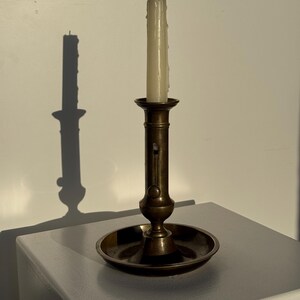 May include: A brass candlestick with a partially burned white candle. The candlestick has a circular base and a long, thin stem. The candle has visible wax drips. The candlestick is on a white surface. A shadow is cast on the wall.
