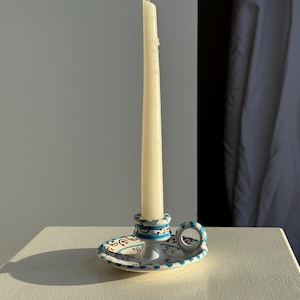 Peut inclure: Un bougeoir en céramique blanche avec une longue bougie crème. Le bougeoir a une base circulaire et une petite poignée, décorée de motifs bleus, rouges et dorés. La bougie mesure environ 20 cm de haut.