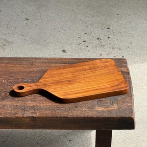 May include: A rectangular wooden chopping board with a smooth, warm brown finish and a prominent wood grain pattern. The board features a short, integrated handle on one side with a circular hole for hanging, showcasing a clean, flat surface.