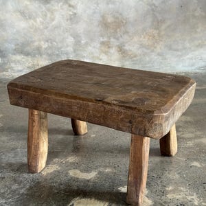 Puede incluir: Un taburete bajo rectangular de madera con un acabado marrón oscuro desgastado. La pieza presenta una superficie gruesa y sólida con vetas visibles y cuatro patas robustas ligeramente cónicas que proporcionan una base estable al mueble.