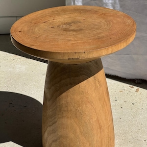 May include: A handcrafted wooden stool with a round top and a conical base. The natural wood grain is visible throughout the stool, showcasing its organic texture. The stool is approximately 18 inches in diameter and 16 inches tall.