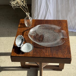 May include: A clear glass fish-shaped serving platter with textured scales rests on a dark wooden table. Two small, shell-shaped dishes and a small glass vase with dried flowers are also on the table. The table has a square top and a cross-legged base.