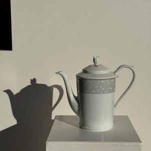 Peut inclure: Une théière en porcelaine blanche avec une bande grise et dorée à motifs sur le dessus. La théière a un long bec et une anse incurvée. Le couvercle est surmonté d'un petit motif floral. La théière est sur une surface blanche.