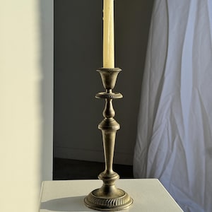 May include: An antique brass candlestick with a cream-coloured candle. The candlestick has a detailed base, a slender stem, and a decorative cup to hold the candle. The background is a neutral white and grey.