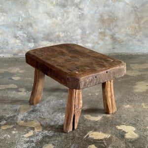 Peut inclure: Un petit tabouret bas en bois doté d'un plateau rectangulaire épais et de quatre pieds robustes légèrement effilés. Le bois présente une finition marron foncé patinée avec un grain visible, des imperfections naturelles et des assemblages apparents.