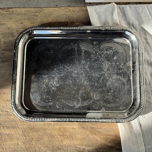 Peut inclure: Un plateau rectangulaire argenté avec un bord perlé et des motifs floraux gravés. Le plateau est posé sur une surface en bois, avec un tissu blanc en arrière-plan. Le plateau est probablement vintage.