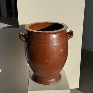 Op de afbeelding: Een bruine keramische pot met twee handvatten aan de zijkant. De pot heeft een brede opening en een afgeronde body die taps toeloopt naar een kleinere basis. De pot wordt tentoongesteld op een witte sokkel. De buitenkant van de pot heeft een glanzende afwerking.
