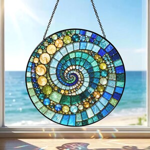 May include: A round, stained glass window hanging with a spiral design. The mosaic features a variety of blue, green, and yellow glass pieces. The window is framed in black and hangs from a chain. The background shows a view of the ocean.
