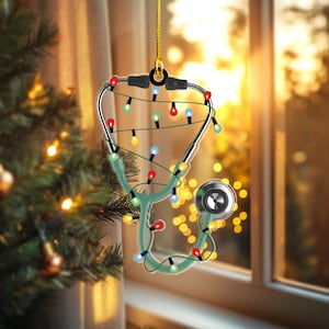 May include: A Christmas ornament shaped like a stethoscope, decorated with colorful string lights. The stethoscope is green and silver, with a gold hanging string. The background is a blurred view of a Christmas tree and a window.