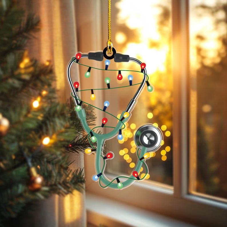 May include: A Christmas ornament shaped like a stethoscope, decorated with colorful string lights. The stethoscope is green and silver, with a gold hanging string. The background is a blurred view of a Christmas tree and a window.