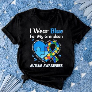 May include: Black t-shirt with the text "I Wear Blue For My Grandson" and "Autism Awareness." The design includes a blue puzzle piece heart, a ribbon, and supporting words like "Family" and "Hope."