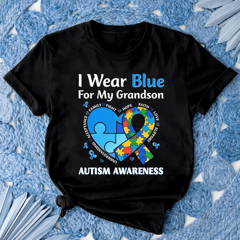 May include: Black t-shirt with the text "I Wear Blue For My Grandson" and "Autism Awareness." The design includes a blue puzzle piece heart, a ribbon, and supporting words like "Family" and "Hope."