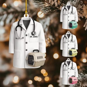 May include: White lab coat ornament with a stethoscope and a small pet carrier. The lab coat has the text "DR. JAMES" on the pocket. The pet carrier is tan and black. The ornament is hanging from a gold string.