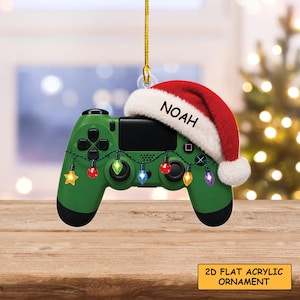 May include: A green video game controller ornament featuring a red and white Santa hat with the name NOAH printed on the brim. A string of colorful Christmas lights is draped across the front of the controller, which hangs from a gold cord.