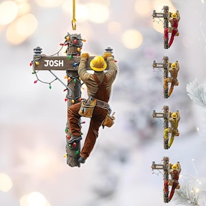 May include: A decorative ornament featuring a lineman climbing a utility pole decorated with Christmas lights. The sign on the pole reads "JOSH". The lineman wears a yellow hard hat, brown overalls, and a tool belt. Additional ornaments with different colored outfits are also shown.