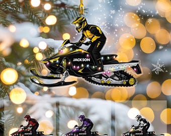 May include: A clear acrylic Christmas ornament featuring a snowmobile with a rider in a yellow and black suit. The snowmobile is black and yellow with the name "JACKSON" on it. Additional ornaments in red, purple, and silver are shown below.