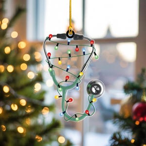 May include: A Christmas ornament shaped like a stethoscope, with a green body and silver earpieces. The stethoscope is decorated with a string of colorful Christmas lights and a gold hanging string. The ornament is displayed in front of a blurred Christmas tree.