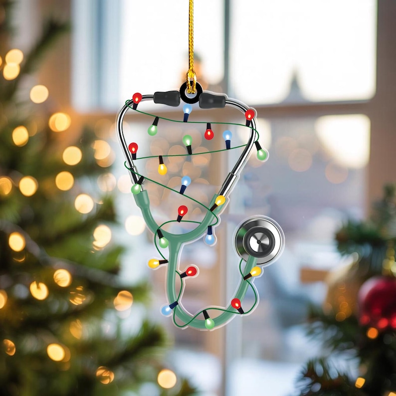 May include: A Christmas ornament shaped like a stethoscope, with a green body and silver earpieces. The stethoscope is decorated with a string of colorful Christmas lights and a gold hanging string. The ornament is displayed in front of a blurred Christmas tree.