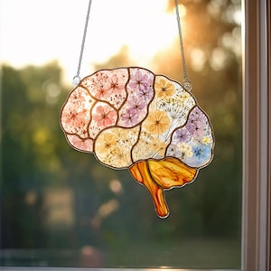 May include: A brain-shaped suncatcher featuring pressed flowers in shades of pink, yellow, purple, and blue encased in clear glass with a copper-toned metal frame. The lower section consists of amber-colored glass, and it hangs from a silver chain.