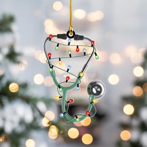 May include: A Christmas ornament shaped like a stethoscope, with a green body and silver accents. The ornament is decorated with colorful Christmas lights and a gold hanging string. The background is blurred with bokeh lights.