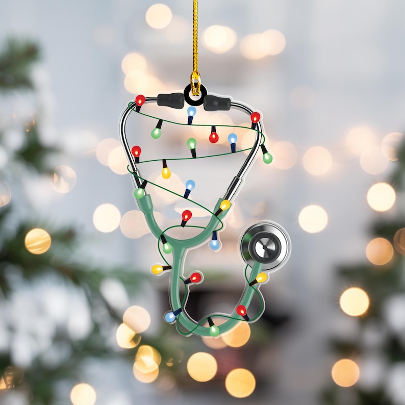 May include: A Christmas ornament shaped like a stethoscope, with a green body and silver accents. The ornament is decorated with colorful Christmas lights and a gold hanging string. The background is blurred with bokeh lights.