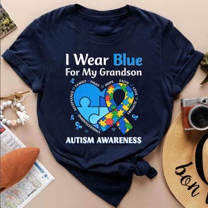 May include: Navy blue t-shirt with the text "I Wear Blue For My Grandson" and "Autism Awareness". The design includes a blue heart-shaped puzzle piece and a colorful ribbon with supporting words.
