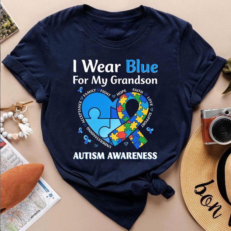 May include: Navy blue t-shirt with the text "I Wear Blue For My Grandson" and "Autism Awareness". The design includes a blue heart-shaped puzzle piece and a colorful ribbon with supporting words.