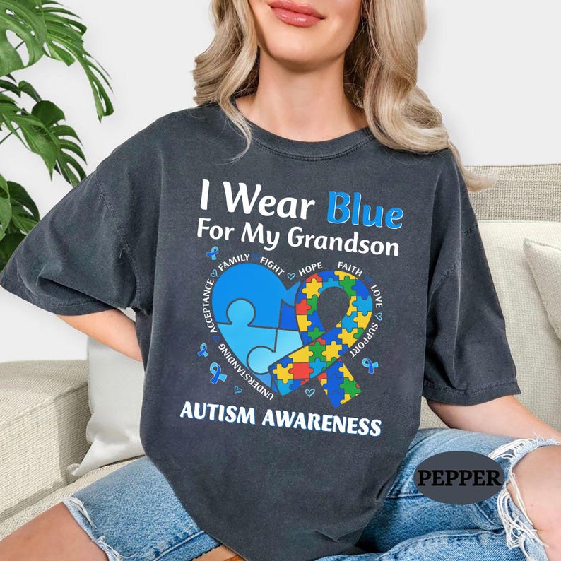 May include: Dark gray t-shirt with the text "I Wear Blue For My Grandson" and "Autism Awareness". The design includes a blue puzzle piece heart and a ribbon with colorful puzzle pieces. The shirt also has the words "Family, Fight, Hope, Faith, Acceptance, Understanding, Support, Love".