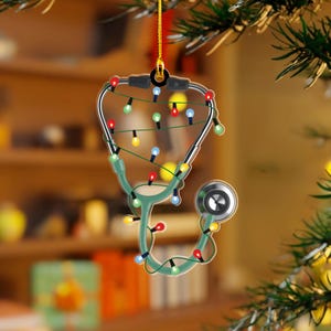 May include: A Christmas tree ornament shaped like a stethoscope, decorated with colorful Christmas lights. The stethoscope is green with silver accents and a gold hanging string. The background is blurred, suggesting a festive setting.