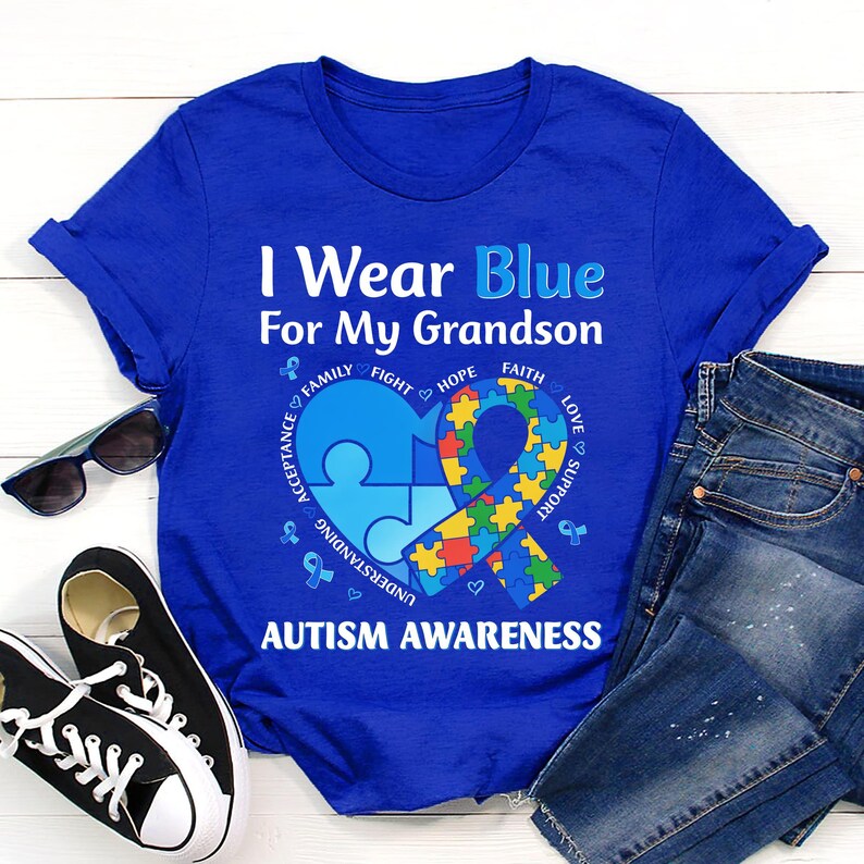 May include: Royal blue t-shirt with the text "I Wear Blue For My Grandson" and "Autism Awareness". The design includes a blue puzzle piece heart and a ribbon with puzzle piece patterns. Black sunglasses, black sneakers, and blue jeans are also visible.
