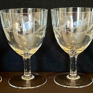 May include: Four clear wine glasses with etched floral designs. Each glass features a rounded bowl, a slender stem, and a circular base. The etched design includes a central flower and surrounding leaves.
