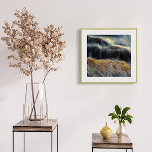 May include: A framed abstract art print with a gold frame. The artwork depicts blurred waves in blue, brown, and gold. The print is displayed on a white wall, with decorative elements including a vase of dried flowers and small side tables.