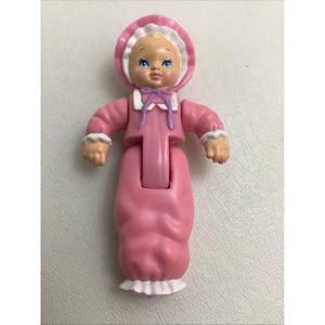 May include: A vintage plastic baby doll in a pink dress and bonnet. The doll has blue eyes, rosy cheeks, and a small purple bow. The dress has white lace trim at the sleeves and hem. The doll's arms are outstretched.