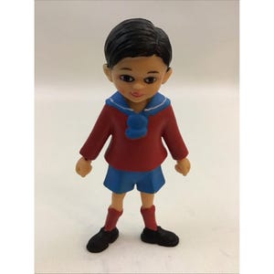 Puede incluir: Una peque&ntilde;a figura de pl&aacute;stico de un ni&ntilde;o que lleva un su&eacute;ter rojo, pantalones cortos azules y zapatos negros. Tiene el pelo negro y un cuello marinero azul.