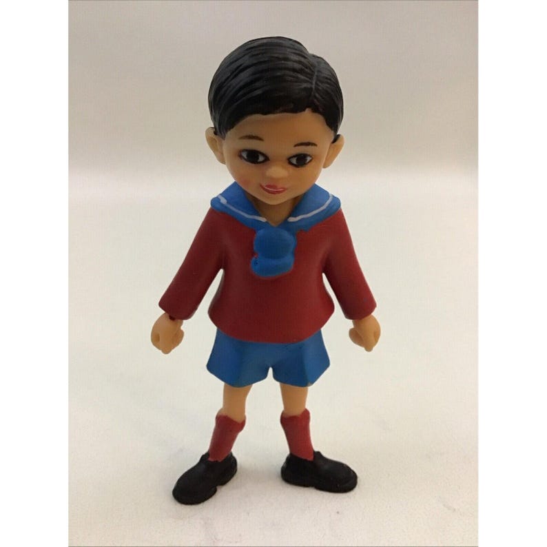 Puede incluir: Una peque&ntilde;a figura de pl&aacute;stico de un ni&ntilde;o que lleva un su&eacute;ter rojo, pantalones cortos azules y zapatos negros. Tiene el pelo negro y un cuello marinero azul.