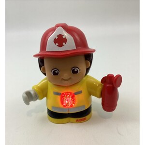 May include: A plastic firefighter figurine wearing a red helmet with a white emblem and a yellow jacket with a glowing red light on the chest. The figure holds a red fire extinguisher in one hand and has the name AIDEN printed in red on the base.