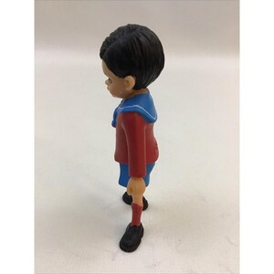 Puede incluir: Una peque&ntilde;a figura de pl&aacute;stico de un ni&ntilde;o que lleva una camisa roja, pantalones cortos azules y un cuello marinero azul y blanco. La figura tiene el pelo negro y est&aacute; de pie con los brazos a los lados.