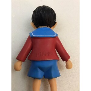 Puede incluir: Una figurita de pl&aacute;stico de un ni&ntilde;o con una camisa de marinero y pantalones cortos, de color rojo y azul. La figurita est&aacute; hecha de pl&aacute;stico y tiene una inscripci&oacute;n blanca en la parte posterior que dice "Pierre".