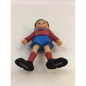 Puede incluir: Una figurita de pl&aacute;stico de un ni&ntilde;o que lleva una camisa roja, pantalones cortos azules y zapatos negros. Los zapatos tienen los n&uacute;meros 443690 y 14799 impresos.