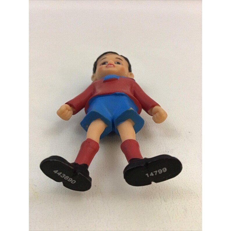 Puede incluir: Una figurita de pl&aacute;stico de un ni&ntilde;o que lleva una camisa roja, pantalones cortos azules y zapatos negros. Los zapatos tienen los n&uacute;meros 443690 y 14799 impresos.