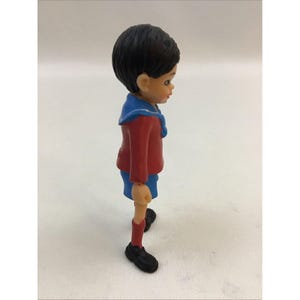 Puede incluir: Una peque&ntilde;a figura de pl&aacute;stico de un ni&ntilde;o que lleva una camisa roja con cuello azul, pantalones cortos azules y zapatos negros. El ni&ntilde;o tiene el pelo negro y la cara pintada.