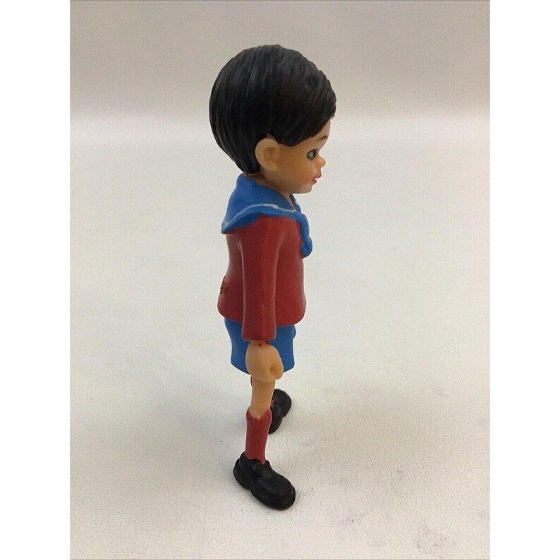 Puede incluir: Una peque&ntilde;a figura de pl&aacute;stico de un ni&ntilde;o que lleva una camisa roja con cuello azul, pantalones cortos azules y zapatos negros. El ni&ntilde;o tiene el pelo negro y la cara pintada.