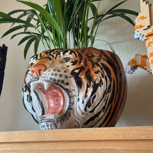 Könnte beinhalten: Ein Keramik-Tigerkopf-Pflanztopf mit offenem Maul, der orange, schwarze und weiße Streifen aufweist. Der Topf enthält eine grüne Blattpflanze. Eine schwarze Katze und eine orange-weiß Katze aus Blöcken befinden sich auf beiden Seiten.
