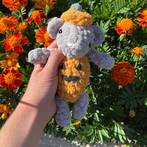 May include: A handmade crochet mouse wearing a pumpkin costume. The mouse is gray with a yellow pumpkin body and hat. The pumpkin has a carved jack-o'-lantern face. The background features orange flowers and green foliage.