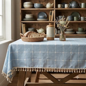 May include: A light blue tablecloth with a white grid pattern and cream-colored tassels along the edges. On the table sits a round woven basket filled with sliced bread, a speckled ceramic French press, and a glass vase containing dried flowers.